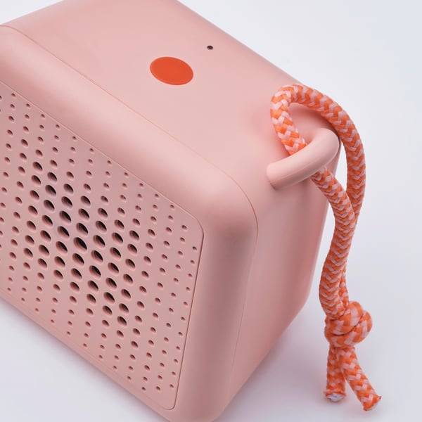 Portable bluetooth speaker in peach with orange handle, speaker grille, and button.