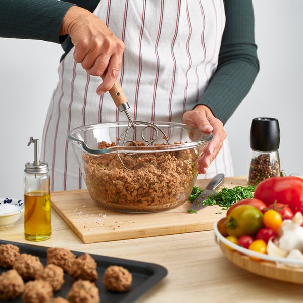 Person mixes minced meat in glass bowl using danish dough whisk. Olive oil, herbs, and tomatoes on wooden table.