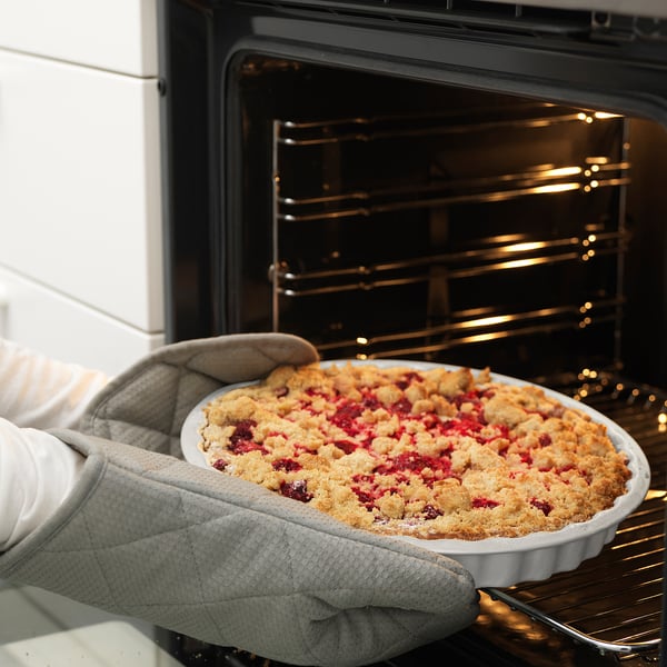 Person holding baked pie in dish near oven. Oven mitts protect hands.