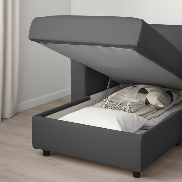 A grey upholstered bed with a lift-up storage compartment at the end, revealing pillows and blankets inside.
