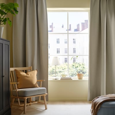 Helles Zimmer mit großen hellgrauen ANNAKAJSA-Vorhängen, einem Holzstuhl und einer Fensterbank, die mit einer Lampe und einer Pflanze geschmückt ist.