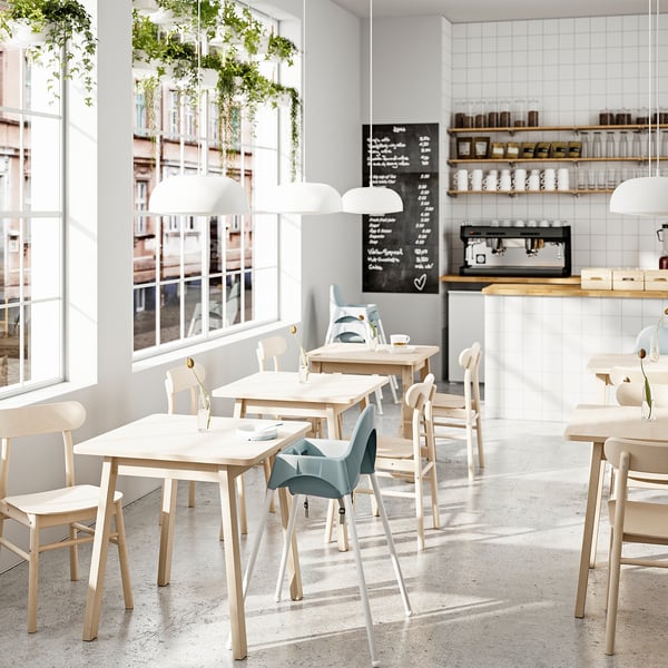 Modernes Café mit hellen Holztischen, Stühlen, großen Fenstern, Hängepflanzen und einer Theke mit Regalen und einer Kaffeemaschine. Helle, luftige Atmosphäre.