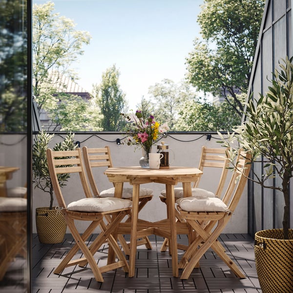 Gemütlicher Balkon mit Holztisch und Stühlen aus ASKHOLMEN , gepolsterten Sitzen, einem floralen Mittelstück und Lichterketten.