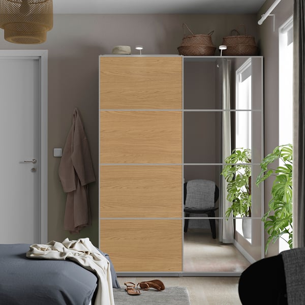 Schlafzimmer mit AULI-Spiegel - und Holzschiebetüren, grauem Bett, Strohhut, Tür, Pflanzen und Stuhl.