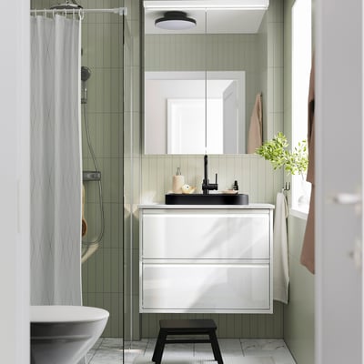 Modernes Badezimmer mit halb eingelassenem Waschbecken aus schwarzem Marmor, weißem Schrank, grünen Fliesen, Glasdusche, Handtuchhaken und Pflanze.