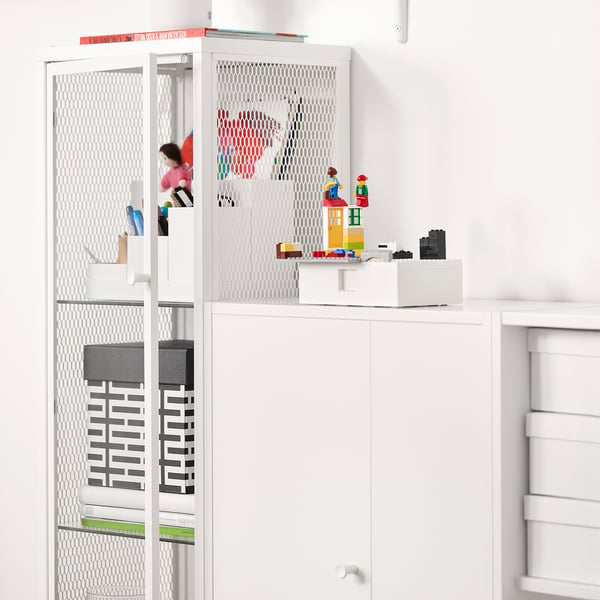 Weißer BAGGEBO-Aufbewahrungsschrank mit Mesh-Paneelen und Glastür mit Lego-Kreationen und farbenfrohen Zeichnungen.