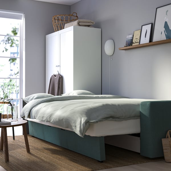 Minimalistisches Schlafzimmer mit BÅRSLÖV-Schlafsofa, das Stauraum und schnellen Bettumbau bietet, vor hellblauer Wand.