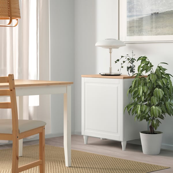 Weißes BESTÅ-Element neben Esstisch mit Pflanze und Lampe, unter großem Fenster.