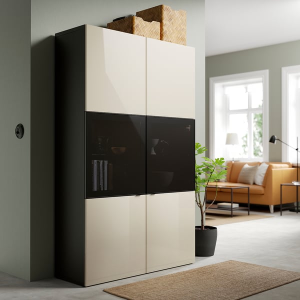 Moderner BESTÅ Aufbewahrungsschrank mit Glastüren, creme und schwarz, im Wohnraum mit Couch und Pflanze.