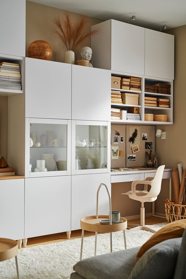 Modernes Home Office mit weißen BESTÅ-Schränken, Glastüren, Büchern, Dekorationsartikeln, Stuhl und kleinem Tisch mit Tasse.