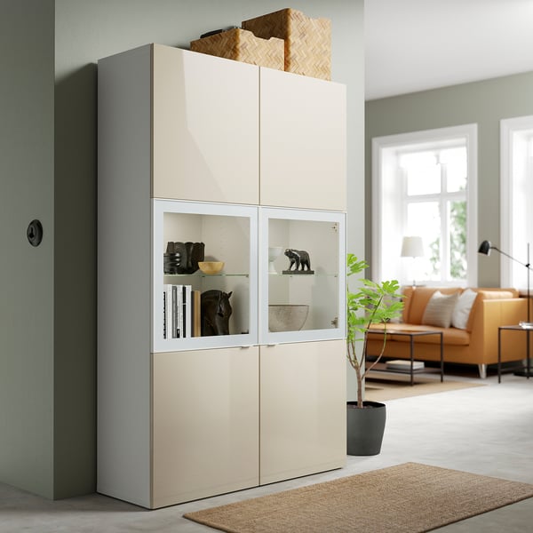 Beigefarbener BESTÅ-Schrank mit grauen Akzenten, hohen, gemischten Regalen und Glastüren.