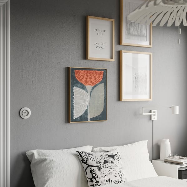Modernes Schlafzimmer mit buntem BLODFLÄDER Poster. Drei gerahmte Drucke an grauer Wand, Bett mit weißen Kissen, Pendelleuchte.