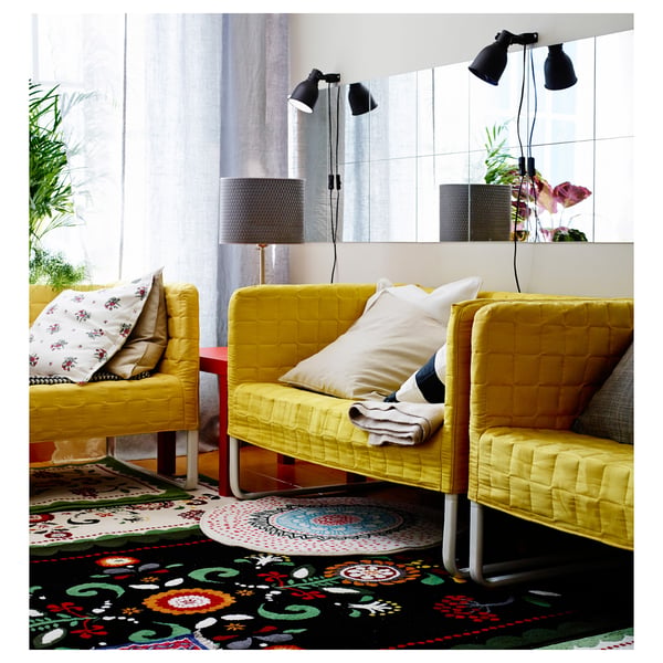 Helles Wohnzimmer mit gelben Stühlen auf einem bunten Teppich, umgeben von Spiegeln und Pflanzen.