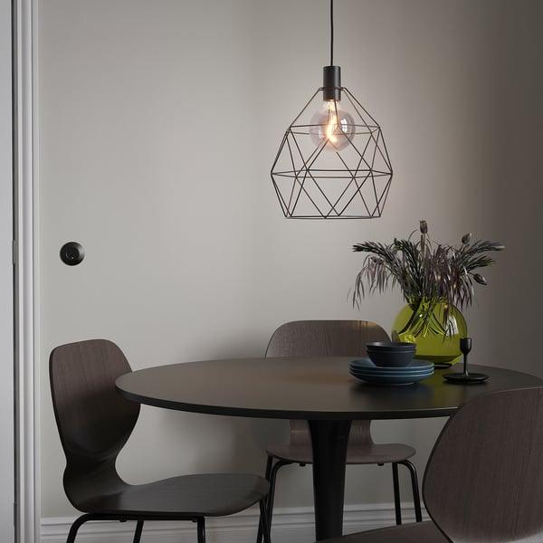 Moderne Esseinrichtung mit BRUNSTA-Lampe, kleinem dunklen Tisch, Holzstühlen, grüner Vase mit Blumen und blauen Tellern.