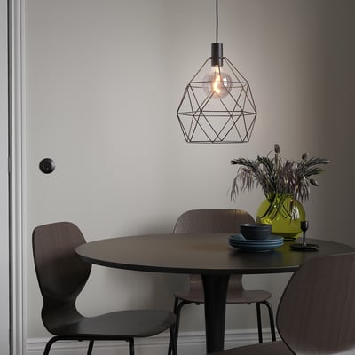 Moderne Esseinrichtung mit BRUNSTA-Lampe, kleinem dunklen Tisch, Holzstühlen, grüner Vase mit Blumen und blauen Tellern.