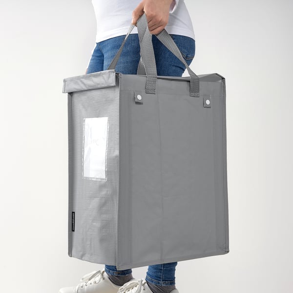Person hält graue DIMPA-Tasche an den Griffen. Die Tasche hat eine durchsichtige Tasche zum Etikettieren, die für die Abfallsortierung geeignet ist.