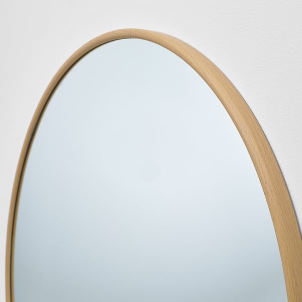 Ein ovaler Spiegel mit dünnem, holzgerahmtem Rand hängt an einer hellen Wand und reflektiert die saubere Oberfläche.