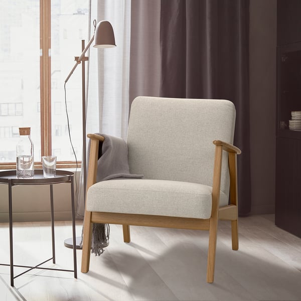 Moderner Stuhl EKENÄSET in neutralen Farben. Holzbeine, weiche Kissen. In der Nähe eines Fensters, mit einem Beistelltisch und einer Lampe. Elegant und komfortabel.