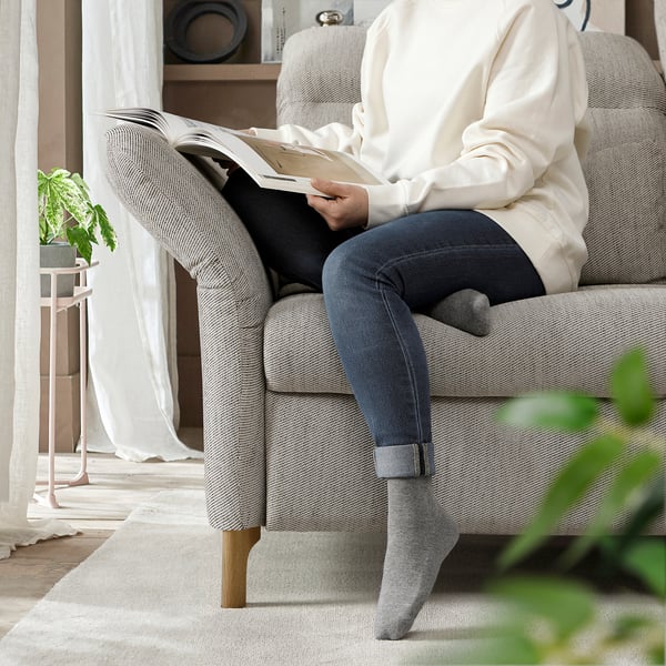 Person liest Buch auf gemütlichem hellem Sofa, Füße gekreuzt, trägt grauen Pullover und Jeans, mit Pflanzen in der Nähe.