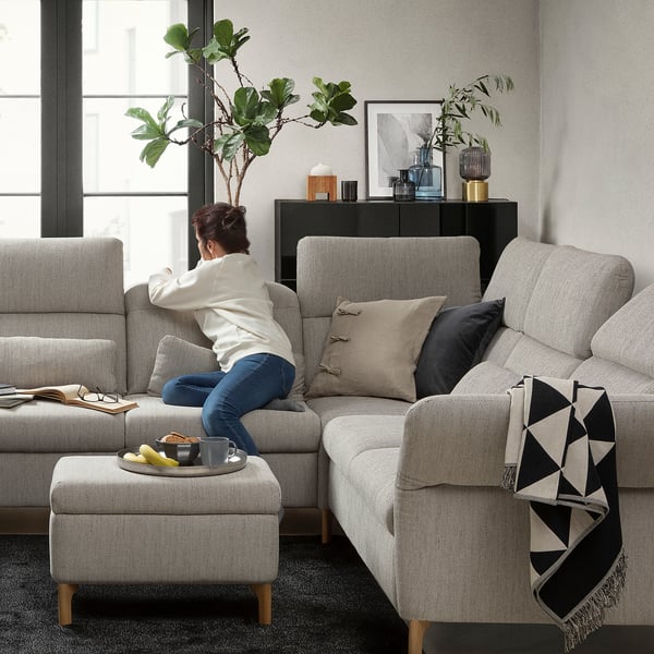 Gemütliches Wohnzimmer mit grauem FAMMARP-Sofa, Kissen und Decke, entspannender Frau, Snacktablett, Pflanzen, Kunst und Fensterblick.