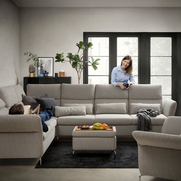 Modernes Wohnzimmer mit hellgrauem FAMMARP-Sektionssofa, verstellbaren Kopfstützen und einer Frau, die ein Gerät benutzt, mit großen Fenstern und Pflanzen.