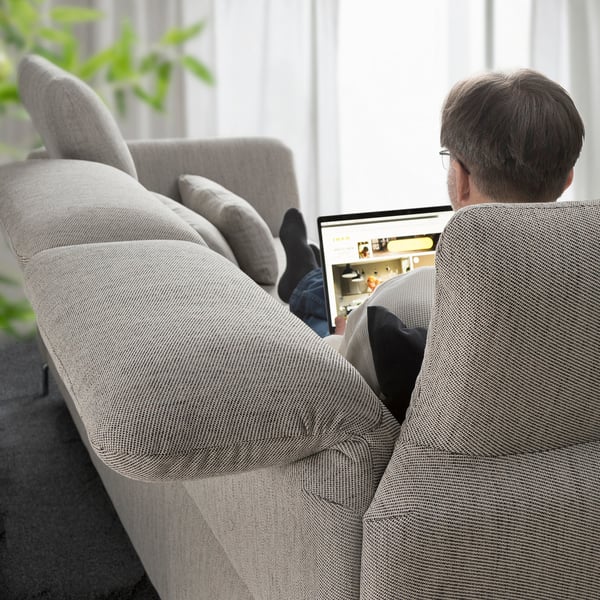 Ein Mann arbeitet an einem Laptop, während er auf einem grauen FAMMARP-Sofa mit verstellbaren Kopfstützen in der Nähe eines Fensters sitzt.