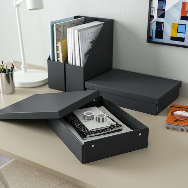 Eine schwarze FJÄDERHARV-Aufbewahrungsbox auf einem Schreibtisch mit Papieren und Notizbüchern, daneben eine Lampe und Kunstdrucke.