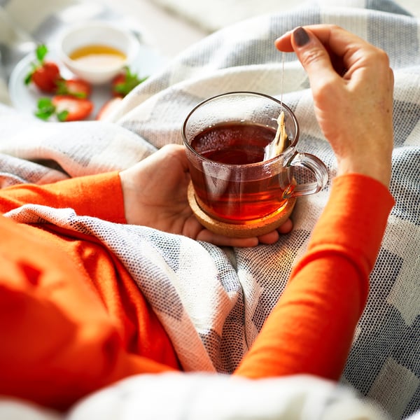 Person mit einem durchsichtigen Glasbecher mit einem Teebeutel, umgeben von kuscheligen Decken und einer Schüssel mit Erdbeeren.
