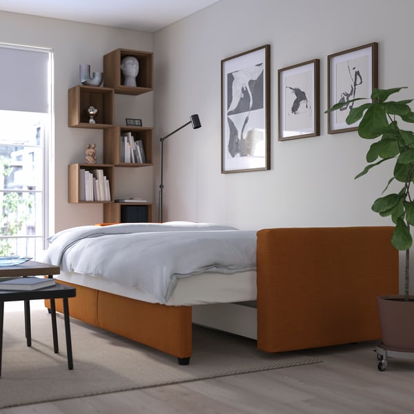 Modernes Schlafzimmer mit ausziehbarem Bett, weißen Bezügen, orangefarbenem Sockel, Pflanze, Bücherregalen und drei Kunstwerken.