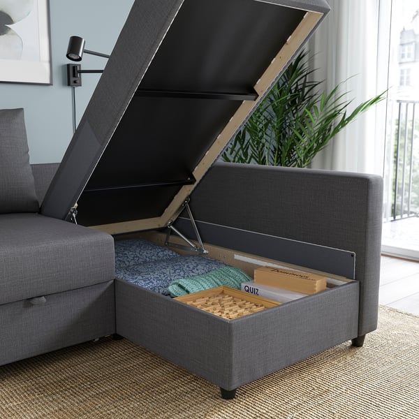 Graues FRIHETEN Schlafsofa mit offenem Stauraum mit Decken und Spielen in einem gut beleuchteten Raum mit Fenster und Pflanze.