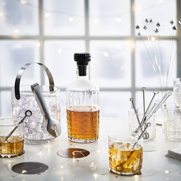 Cocktail-Setup: Klarglasflasche mit schwarzem Verschluss, Eiskübel, Zange, Gläser, Rührstäbchen auf weißem Tisch.
