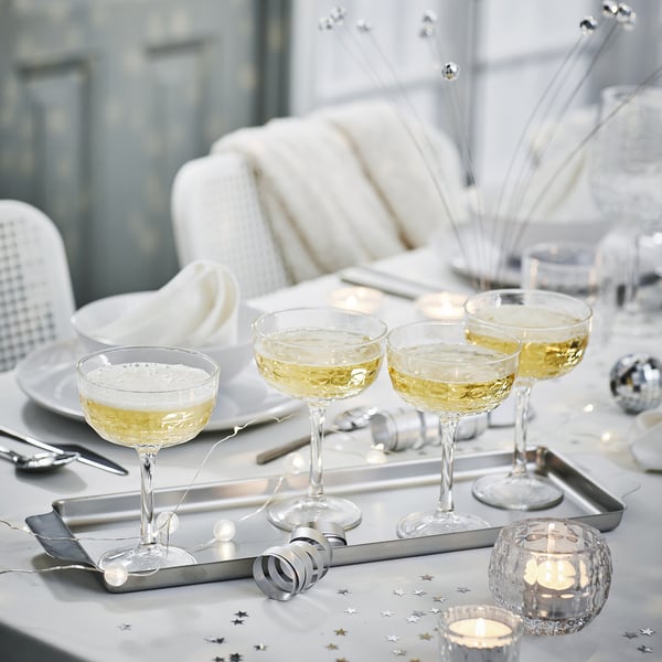 Tischgarnitur mit durchsichtigen FRÖJDA-Champagner-Coupes, silbernem Tablett, Sternkonfetti, hellblauem Hintergrund, weißer Couch, eleganter Einrichtung.