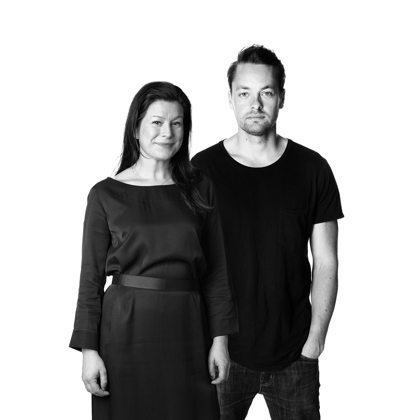 Schwarz-Weiß-Foto des Designers in einem dunklen Kleid und des Designers in einem dunklen T-Shirt, der Seite an Seite steht und lächelt.