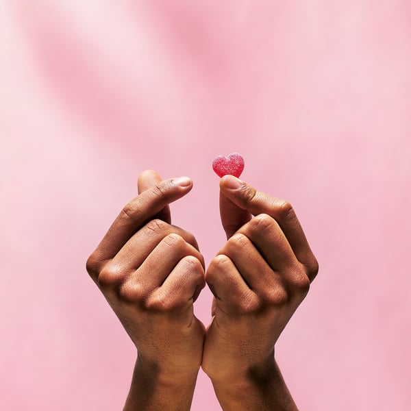 Zwei Hände halten kleine, glitzernde rote, herzförmige Bonbons zwischen den Fingern vor einem rosa Hintergrund.