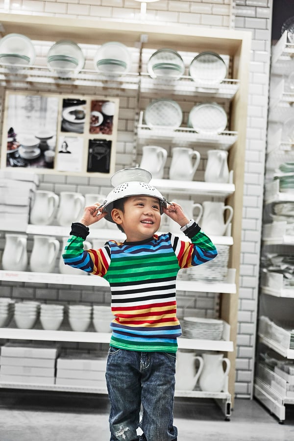 Junger Junge mit bunt gestreiftem Hemd, lachend und mit einem silbernen Bratpfannenhut im Küchengeschäft.