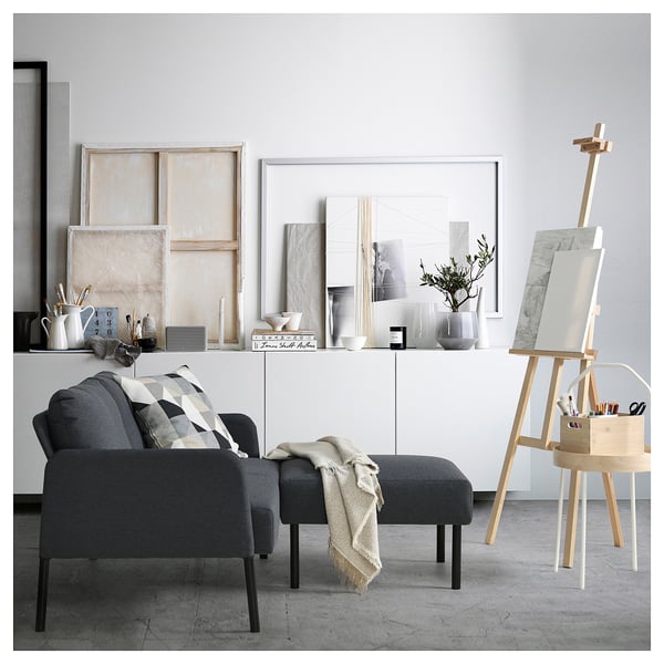 Moderner Wohnbereich mit dunkelgrauem GLOSTAD-Sofa und Ottomane, dekoriert mit minimalistischer Kunst, Vasen und einer Staffelei.