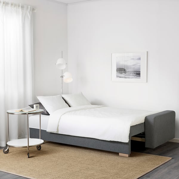 Modernes Schlafzimmer mit GRÄLVIKEN-Schlafsofa, weißer Bettwäsche, Beistelltisch und Stranddruck.