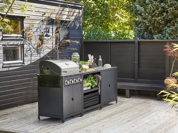 Moderne Outdoor-Küche mit schwarzer GRILLSKÄR-Grillstation mit Edelstahlgeräten, Holzterrasse und dunklem Zaunhintergrund.