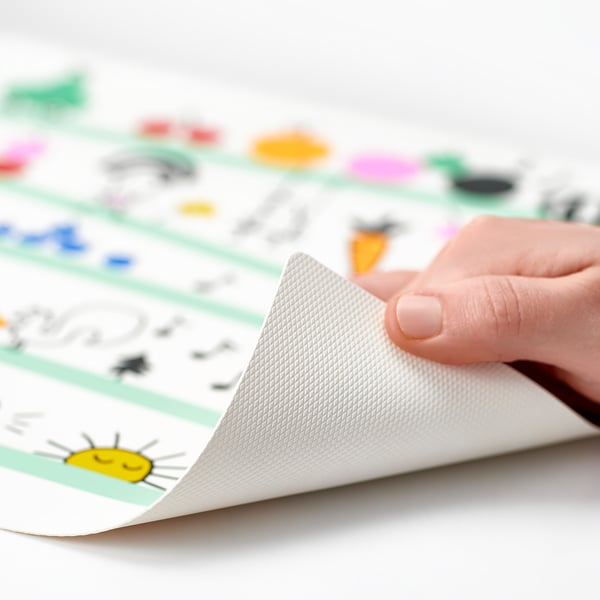 Die Hand schält die Ecke des bunten GRÖNFINK-Tischsets zurück und enthüllt das Griffmuster, das für Kinder entwickelt wurde und die Tische vor Verschütten schützt.