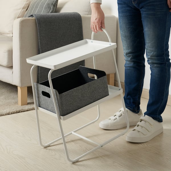 Weißer Beistelltisch mit Regalen und Mülleimer, neben beigem Stuhl. Eine Person steht in der Nähe und zeigt Mobilität.