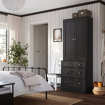 Schlafzimmer mit dunklem HAVSTA-Schrank, weißem Stuhl, schwarzem Bett, Teppich und Pflanze.