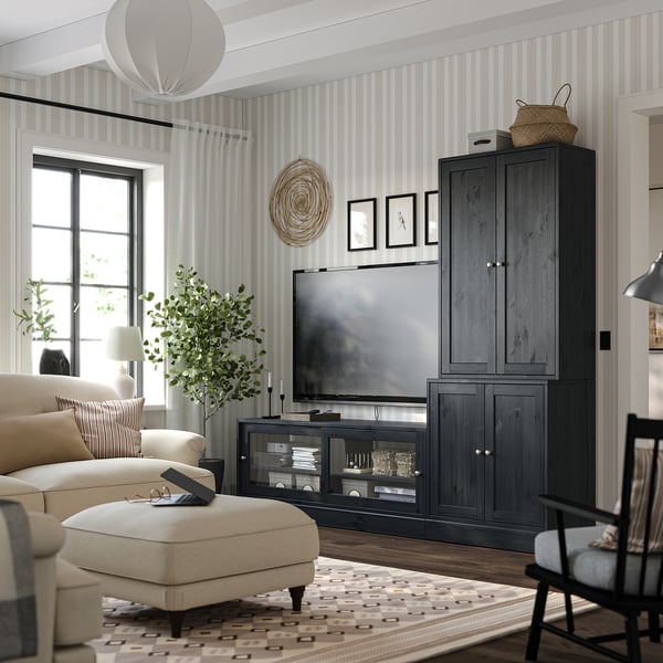 Modernes Wohnzimmer mit gestreifter Tapete, beiger Couch, gemustertem Teppich, TV-Schrank aus dunklem Holz, großem Flachbildfernseher, stilvoller Lampe und einer Pflanze.