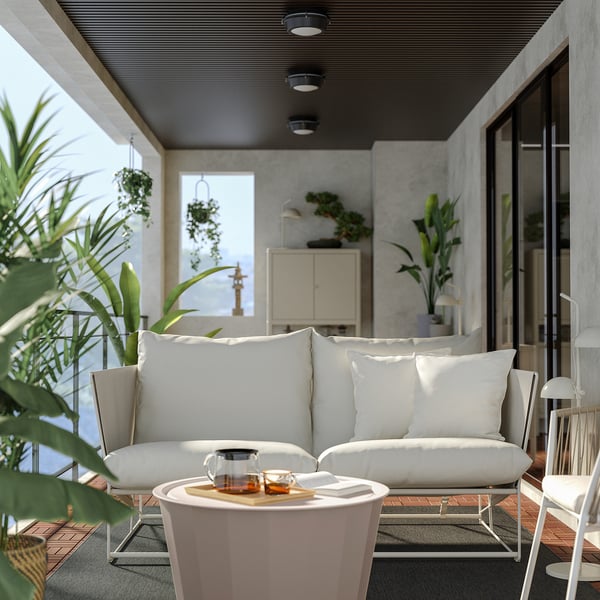 Moderner weißer Innenhof mit HAVSTEN-Sofa, Kissen, Teeservice aus Glas, umgeben von Pflanzen, elegantes Dekor.
