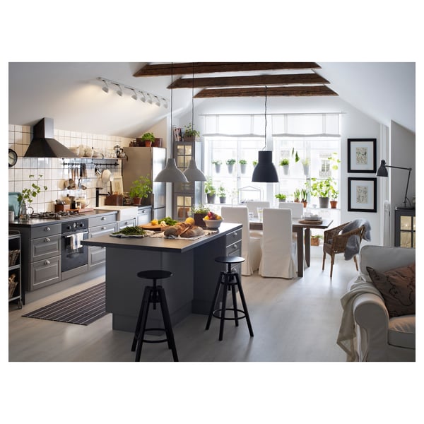 Moderne Küche mit Fabriklampen über Esstisch, Holzbalken, weißen Wänden, grauen Schränken, gemütlichem Wohnbereich mit Sofa, Korbstuhl und Pflanzen.