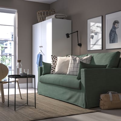 Modernes Wohnzimmer mit grünem Sofa, weißem Schrank, Kunst. Bunte Kissen, Vase, großes Fenster.
