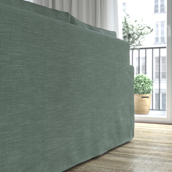 Ein grünes Stoffsofa mit wendbarem Kissen in der Nähe eines Fensters mit Pflanzen, das Komfort und Langlebigkeit bietet.