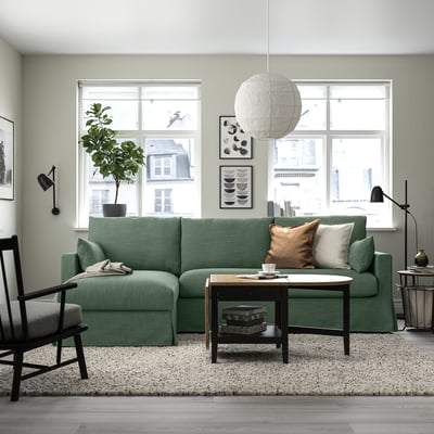 Ein stilvolles Wohnzimmer mit einem grünen HYLTARP-Sofa, einem Couchtisch, einem schwarzen Sessel und einem großen Fenster.