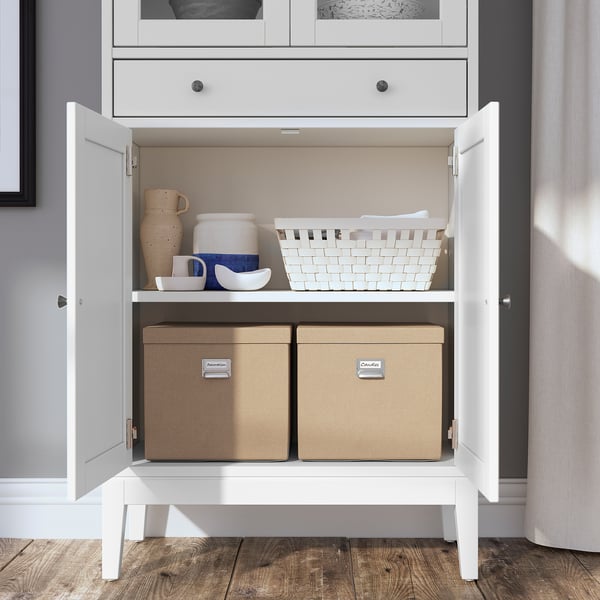 Weißer Schrank mit Glastüren, in dem Keramik und Aufbewahrungsboxen ausgestellt sind. Modernes, funktionales Design.