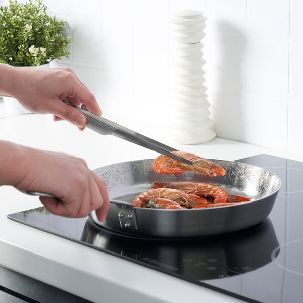 Eine Person benutzt eine Kochpinzette, um Garnelen in einer Pfanne auf einem Herd zu handhaben. Die Pinzette bietet einen festen Halt beim Kochen.