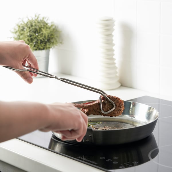 Hände, die mit einer Zange das Steak in einer Pfanne wenden, demonstrieren den Griff von Küchenutensilien und die Kochhilfe.